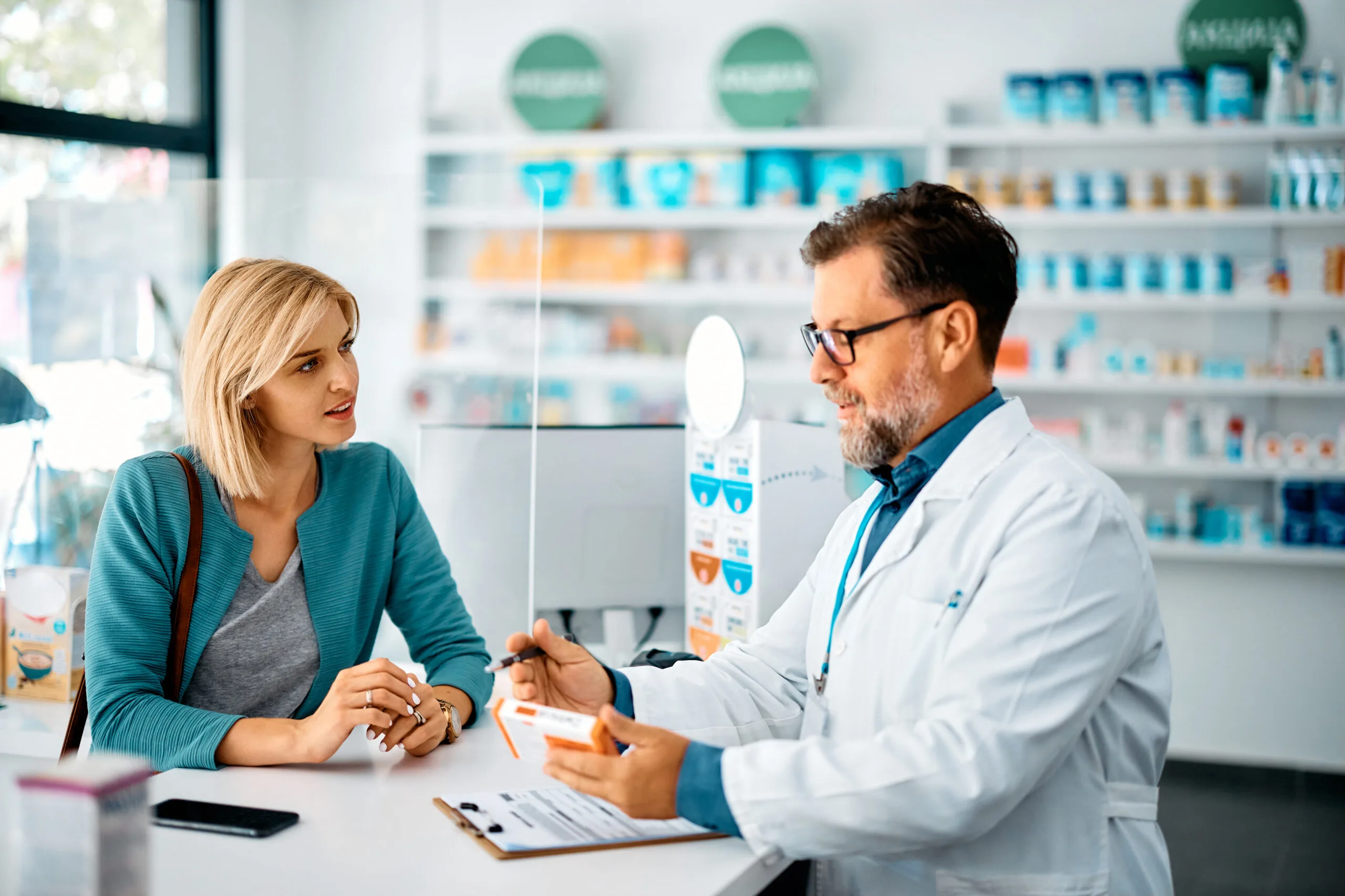 A pharmacist in a white lab coat is attentively explaining a prescription medication to a customer in a bright and well-organized pharmacy. The clear interaction highlights the personalized care and professional advice offered at Edgemont Pharmacy Calgary NW.