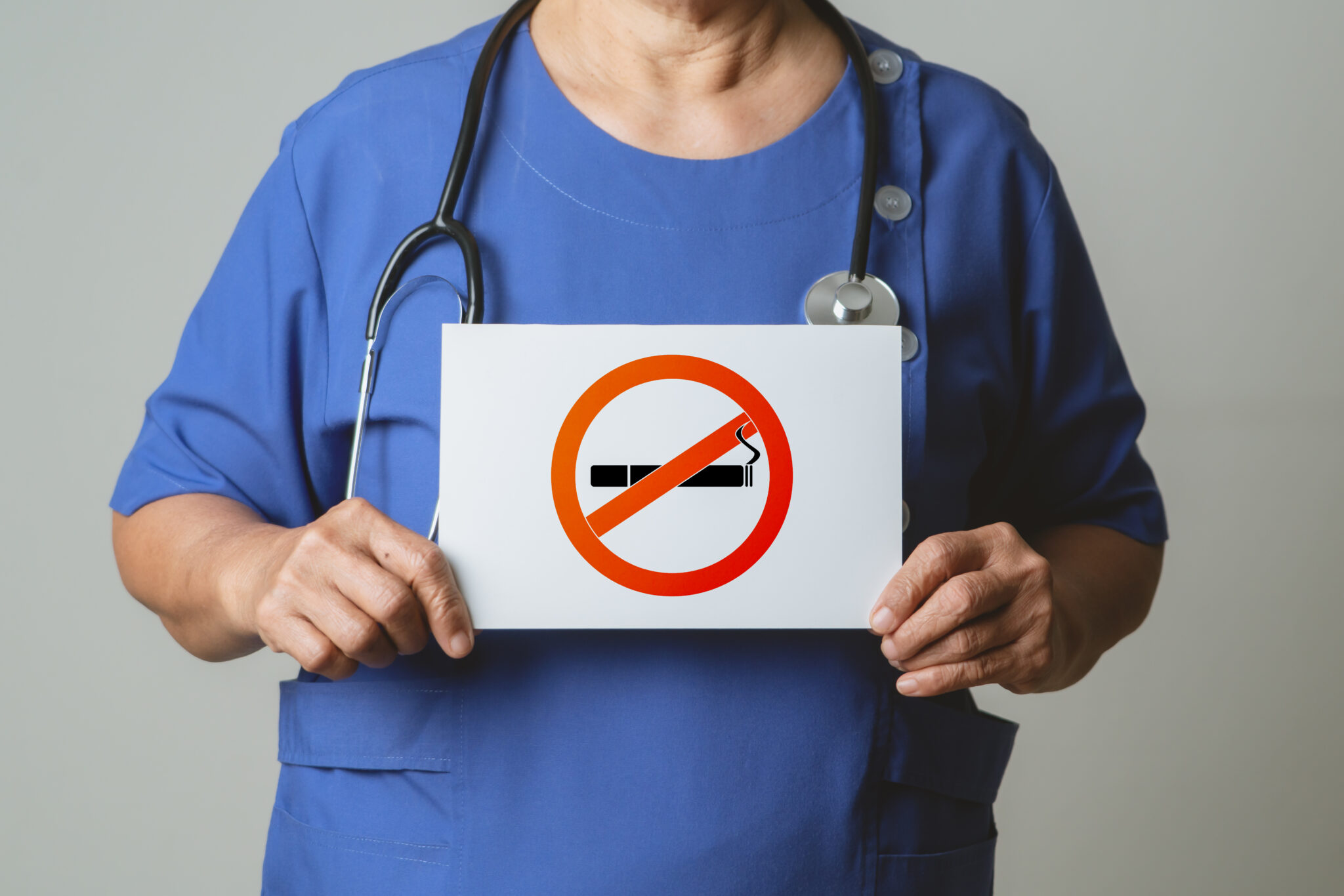 A healthcare professional in blue scrubs and a stethoscope around the neck is holding a sign with the no smoking symbol.