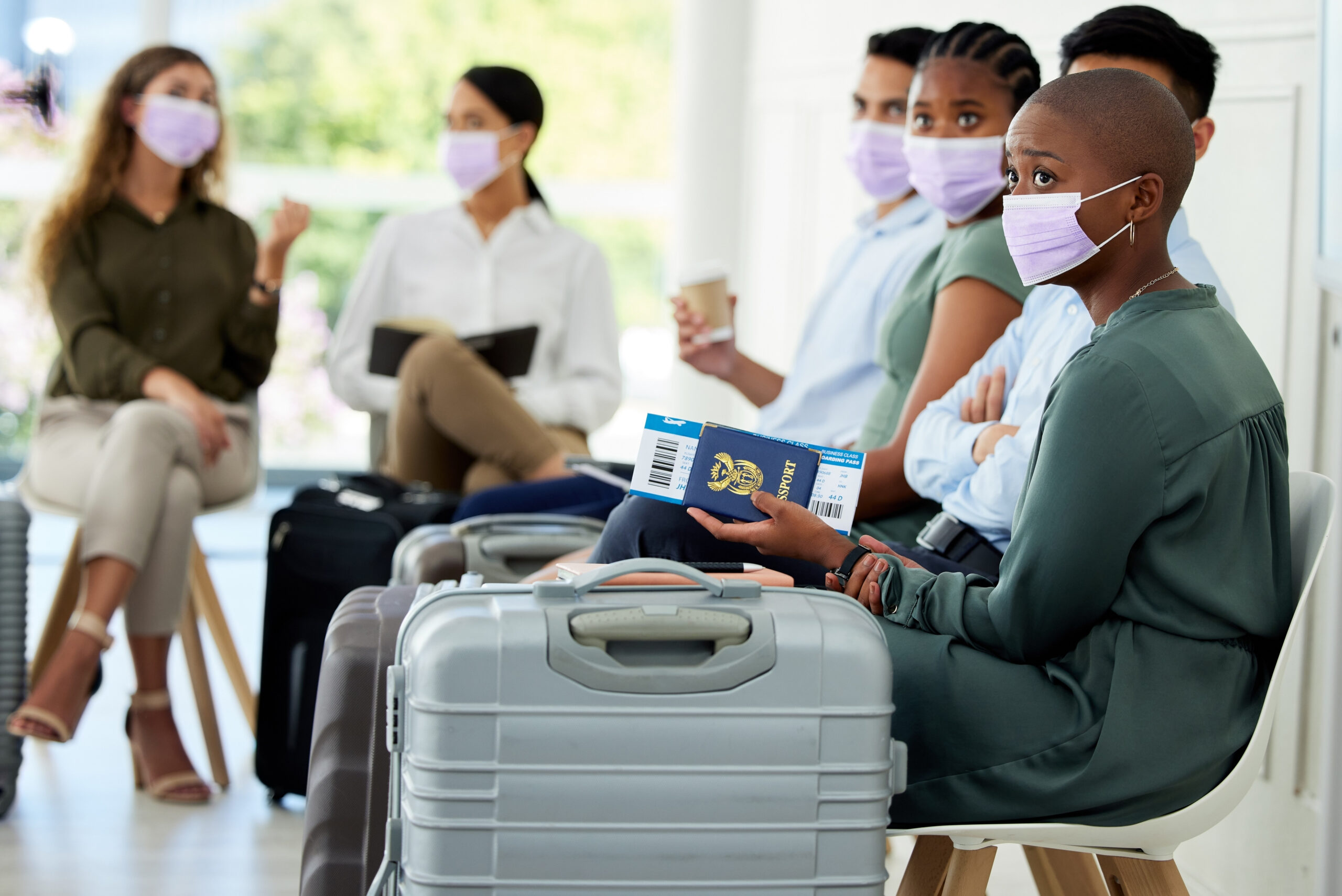 A group of individuals seated in a waiting area, with one person holding a passport and sitting next to a grey suitcase, possibly indicating preparation for travel.