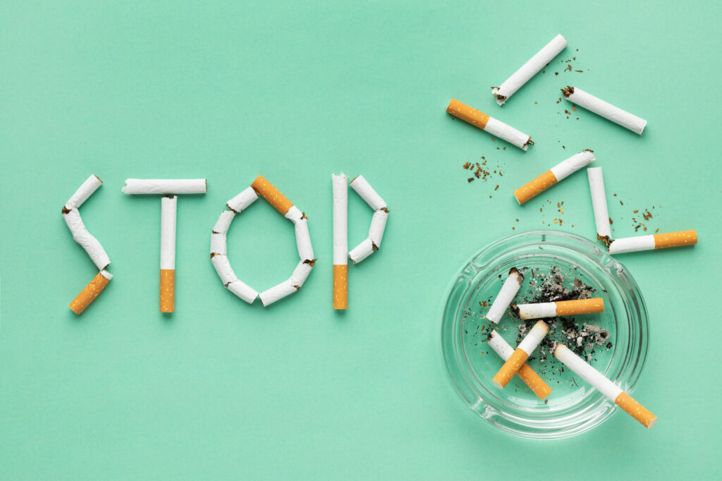 The image shows the word ‘STOP’ spelled out with broken cigarette pieces on a green background, with whole cigarettes and ash scattered around. A clear glass ashtray filled with cigarette butts is also visible.