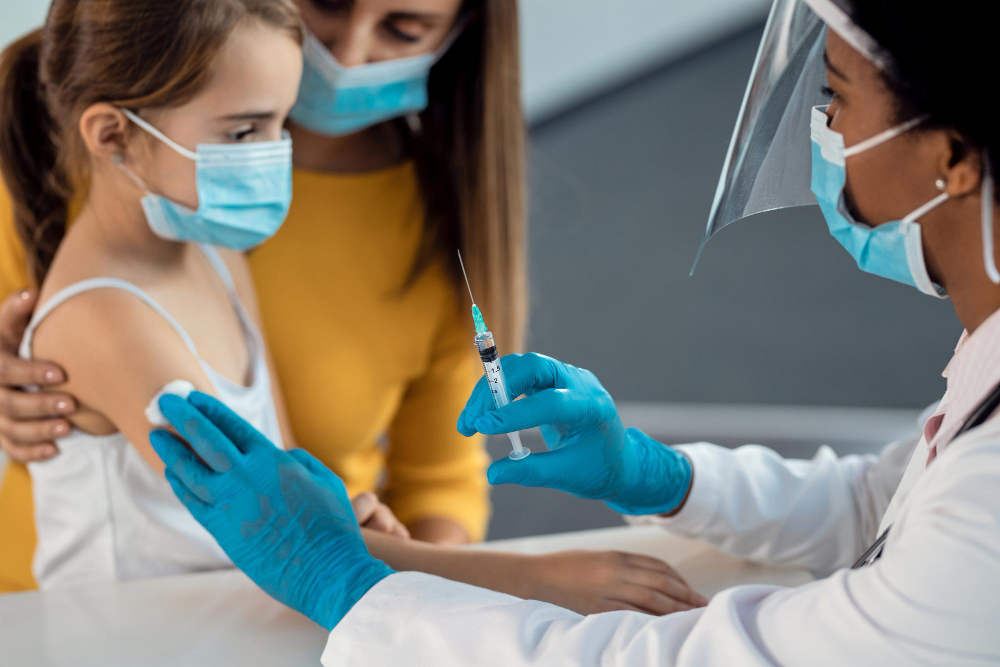 A healthcare professional wearing a face shield and blue gloves administers a vaccine to a young girl, who is comforted by a masked adult.