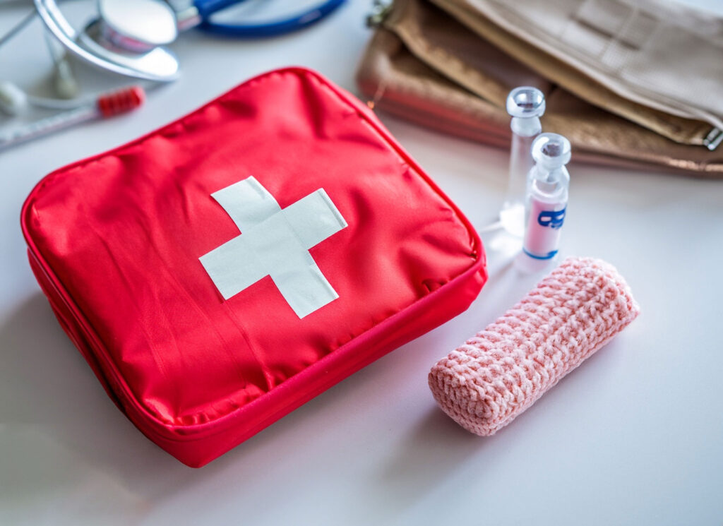 A red first aid kit with a white cross on the top is prominently displayed on a white surface. To the right of the kit, there’s a clear bottle with a blue cap, possibly containing a disinfectant or medication. In the background, there’s a blurred brown bag, suggesting additional medical supplies or personal belongings.