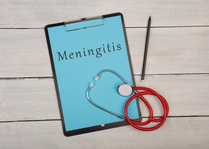 A clipboard with a paper titled ‘Meningitis’ attached to it, resting on a wooden surface. A stethoscope with red tubing is draped over the bottom of the clipboard, and a black pen lies to the right side.